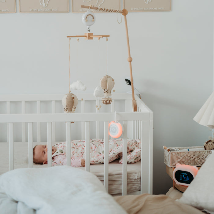 Wide photo with baby in cot, Sound Sleeper portable white noise with Sleep Like Goldilocks smart thermometer in background, both with red light